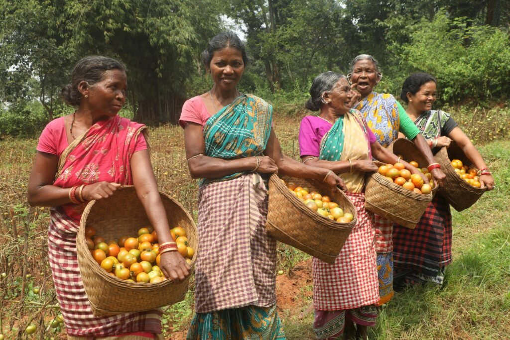 Two smiling women farmers carrying overflowing baskets of fresh fruits on their heads, walking through a lush field in a sustainable agriculture harvest scene